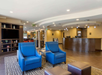 Hotel lobby with blue chairs, a round reception desk, and wall-mounted shelves. Overhead lighting illuminates the space, featuring a neutral color palette and modern design.