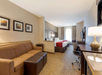 Hotel room with two beds, a sofa, a desk, and a TV. Tan and white color scheme. Artwork on the wall. A window with curtains is in the background.