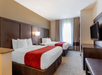 Hotel room with two double beds, each with white linens and a red runner. There is a wall-mounted TV, a window with curtains, and a small table with a lamp between the beds.