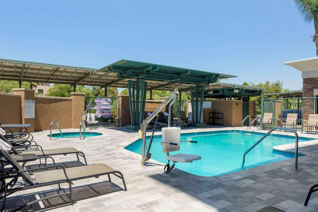 Outdoor pool area with a small pool, lounge chairs, and green pergolas. A mechanical lift and a hot tub are adjacent to the pool. Palm trees and a clear sky are in the background.