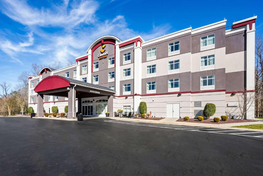 A four-story hotel building with a red and gray facade, a covered entrance, and a parking lot in the foreground on a clear day.