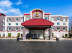 Hotel exterior with a red entrance canopy, multiple windows, and the "Comfort Suites" logo on top. The sky is clear and blue.