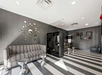 Modern lounge with a striped floor, gray couches, round metal side tables, and decorative wall mirrors. Black shelving unit displays vases and books. Ceiling lights illuminate the space.