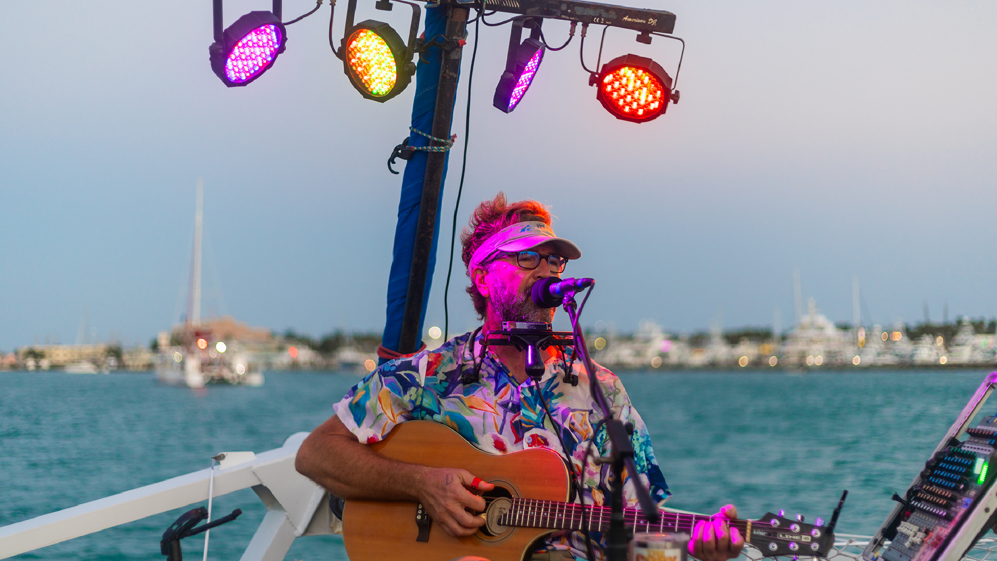 A man wearing a colorful shirt and visor sings and plays guitar on a boat at sunset, with stage lights and a marina in the background.