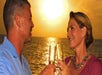 A man and woman toast with champagne glasses, smiling at each other with a sunset over the ocean in the background.