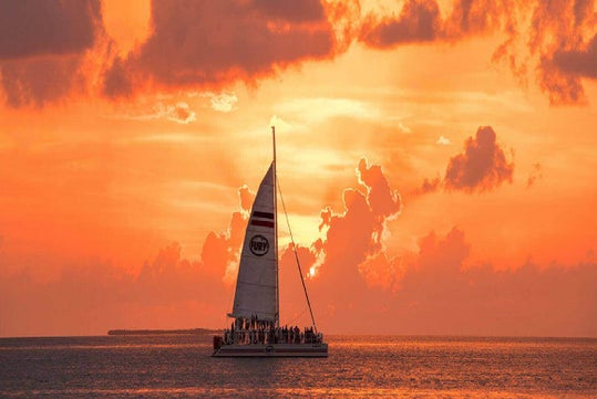 A catamaran sails on the ocean under an orange sunset sky, with clouds scattered above and a silhouette of people on board