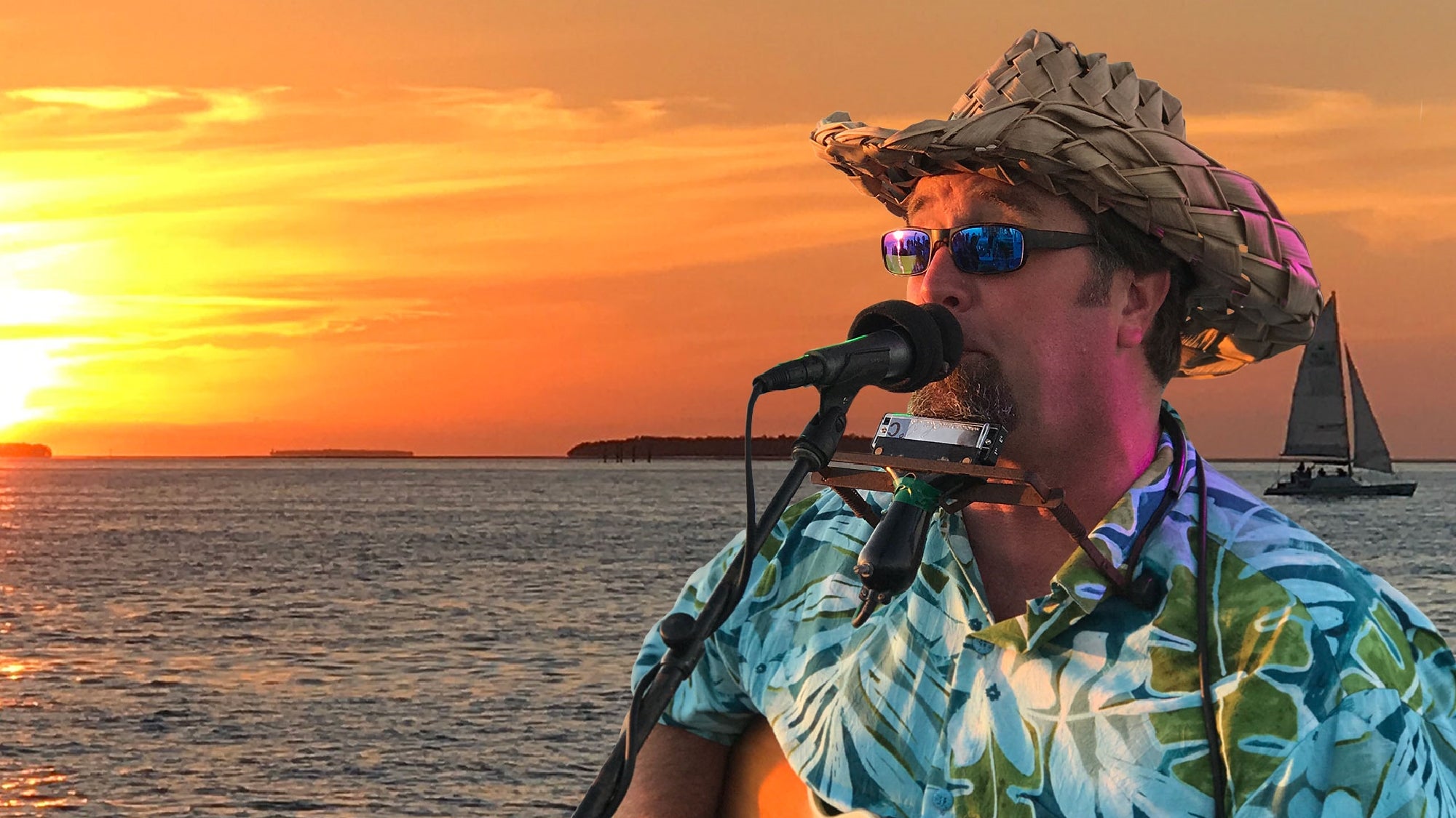 A man with a straw hat and sunglasses sings into a microphone with a sunset and sailboat in the background. He is wearing a tropical shirt and has a harmonica holder around his neck.