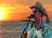 A man in a straw hat and sunglasses sings and plays guitar with a harmonica holder at sunset by the ocean; a sailboat is in the background.