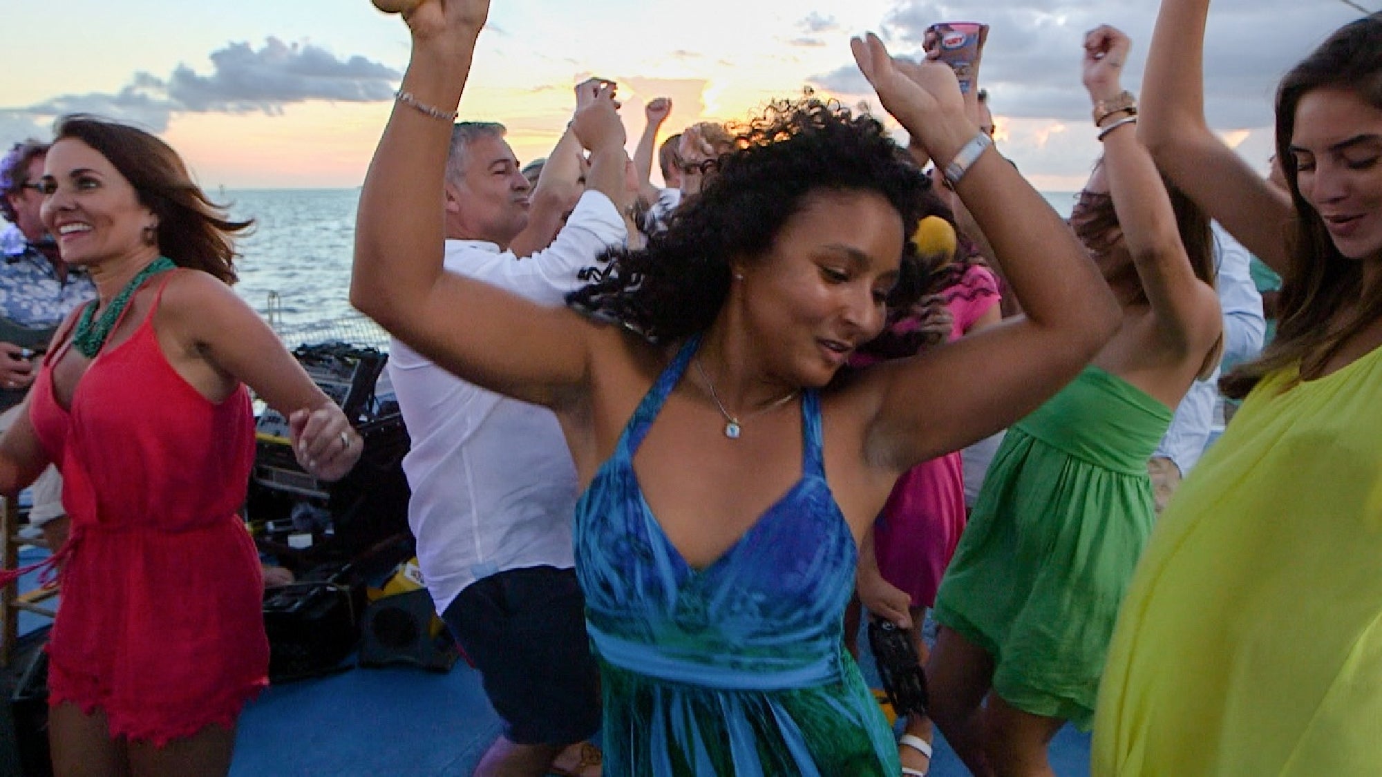 A group of people dance energetically on a boat at sunset, with the ocean in the background. They are dressed in colorful summer outfits