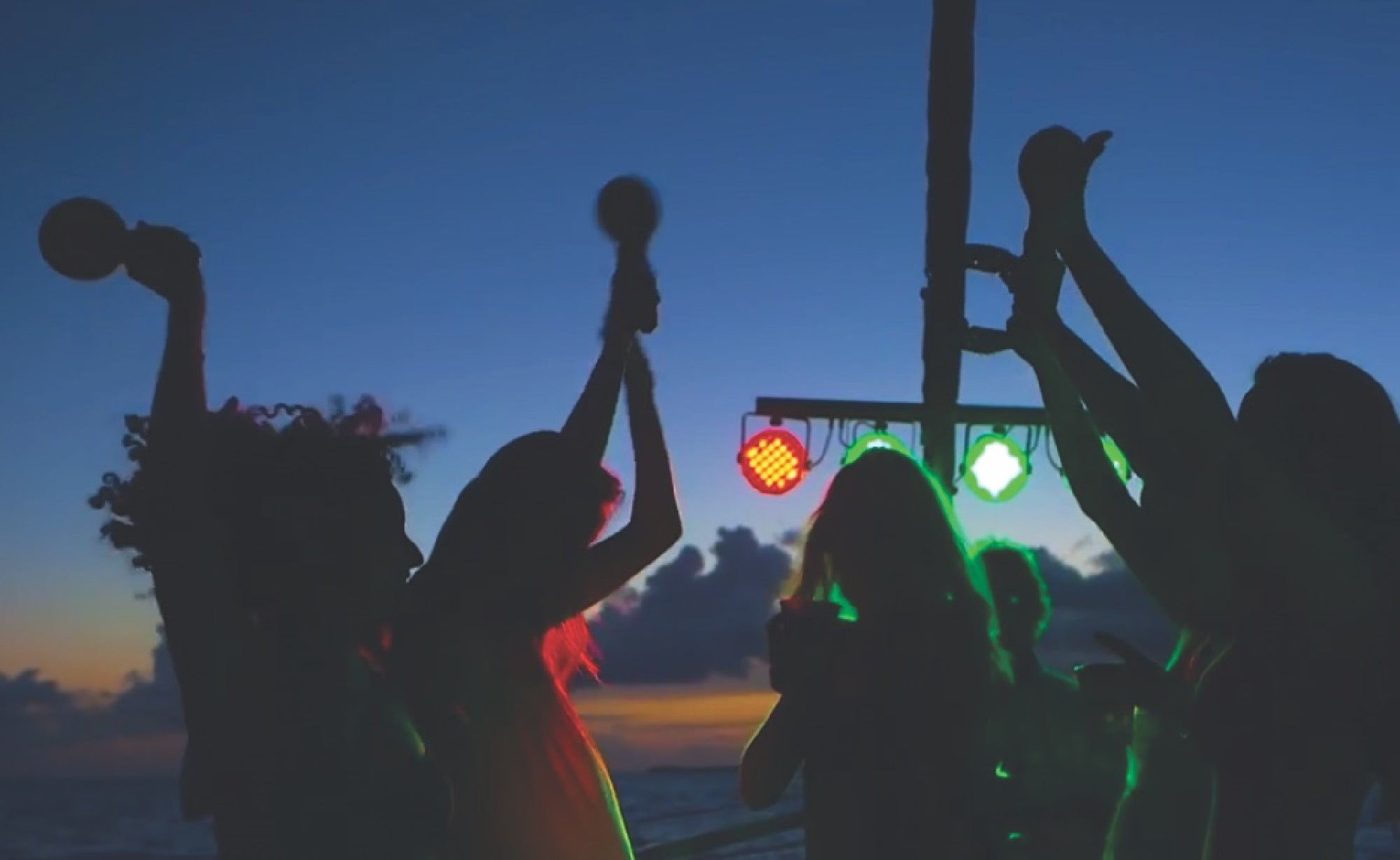 Silhouette of a group of people dancing with maracas in hand against a sunset backdrop with colorful lights.