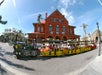 A colorful tourist train with passengers drives past a large red brick building under a blue sky with palm trees in the background.