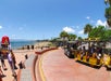 Tourist tram with passengers parked near a waterfront promenade on a sunny day, with people walking and the ocean in the background.