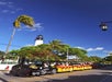 A small tourist train drives down a sunny street lined with palm trees, with a tall lighthouse and blue sky in the background.