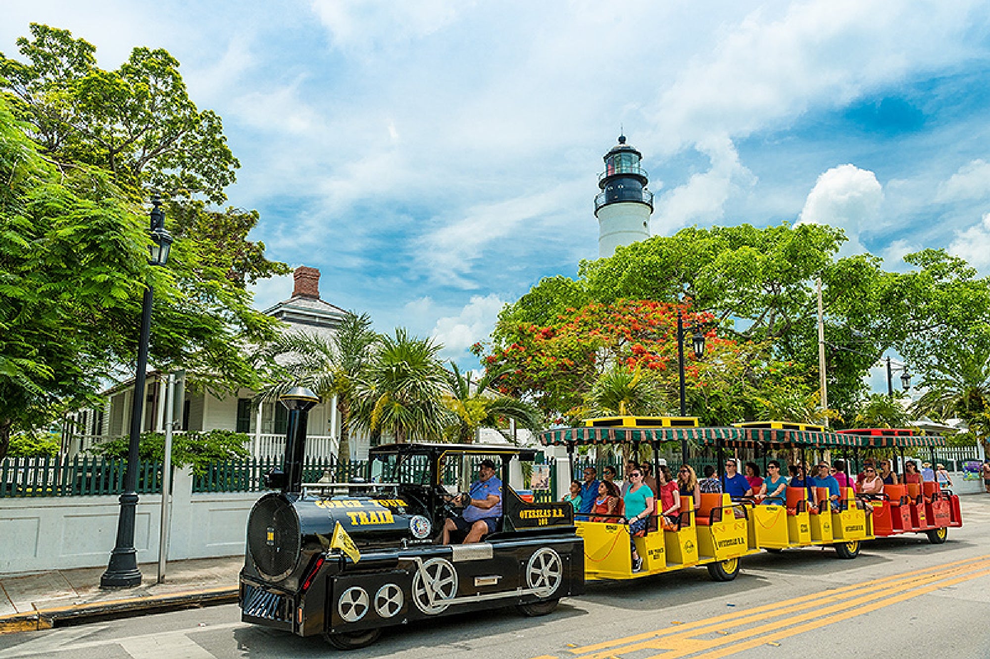A sightseeing tour tram with passengers drives past tropical trees and a white lighthouse under a blue sky with clouds.
