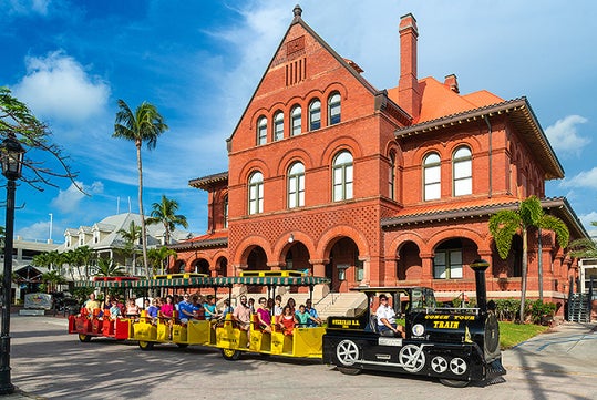 A group of people ride an open-air tour train in front of a large red brick historic building with arched windows and palm trees nearby.
