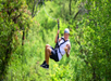 Person wearing a helmet and harness ziplining through a lush green forest, smiling and gesturing with one hand.