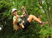 A person wearing a helmet and harness rides a zip line outdoors, surrounded by green trees, and smiles while gesturing a thumbs up.