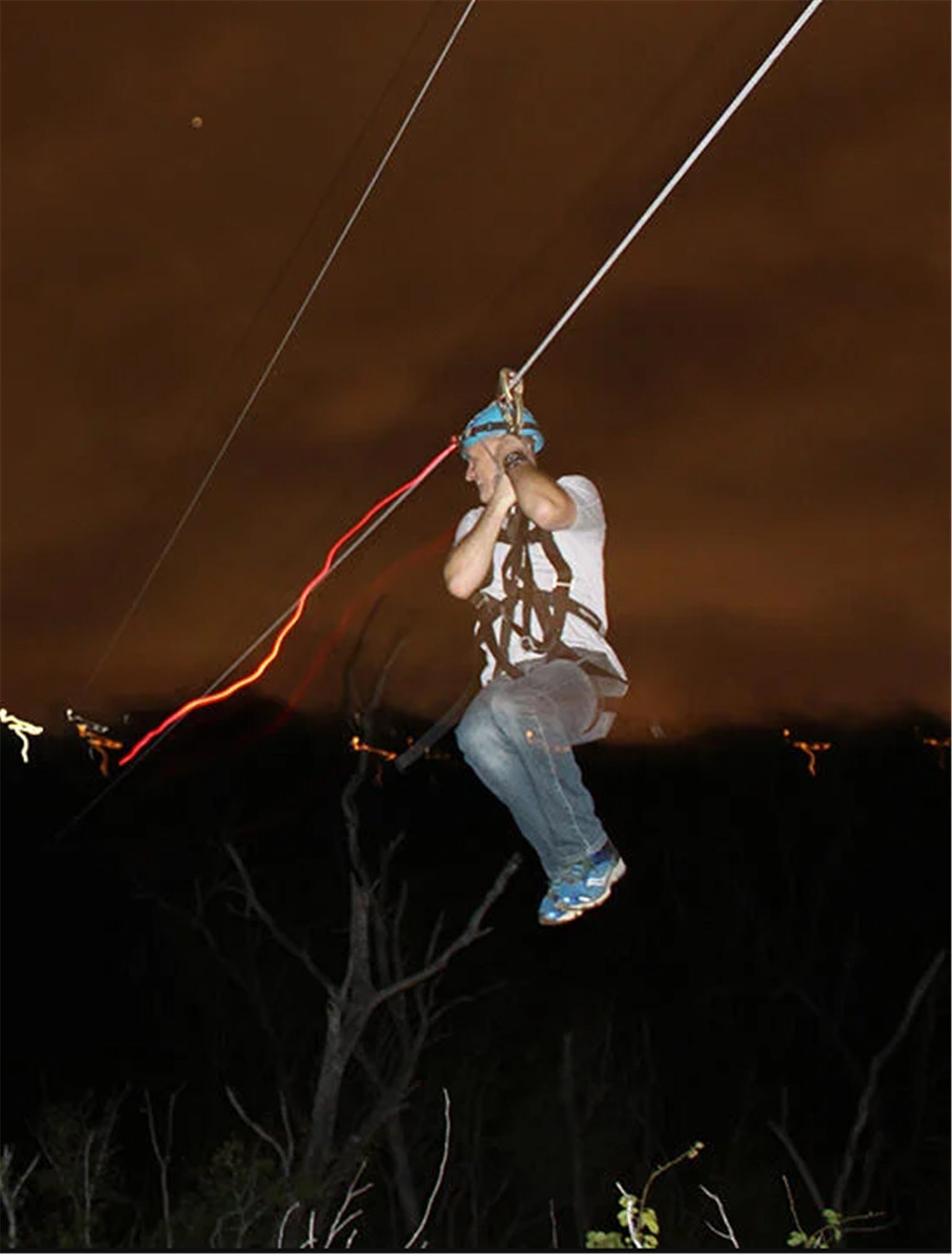 A person wearing a helmet and harness rides a zip line at night, with dark trees and a cloudy sky in the background.
