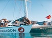 A woman sits on the bow of a sailboat named Paragon, with other people on deck. The boat is anchored on calm water under a clear sky.