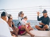 A group of people, including a child, sit on a boat while a man gestures as he speaks. The ocean and clear sky are visible in the background.
