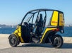 A yellow and black three-wheeled electric vehicle is parked by the waterfront, featuring an open design with two seats and clear skies in the background.