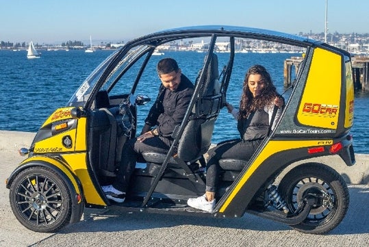 Two people sit in a yellow and black three-wheeled vehicle by the waterfront, with sailboats visible in the background.