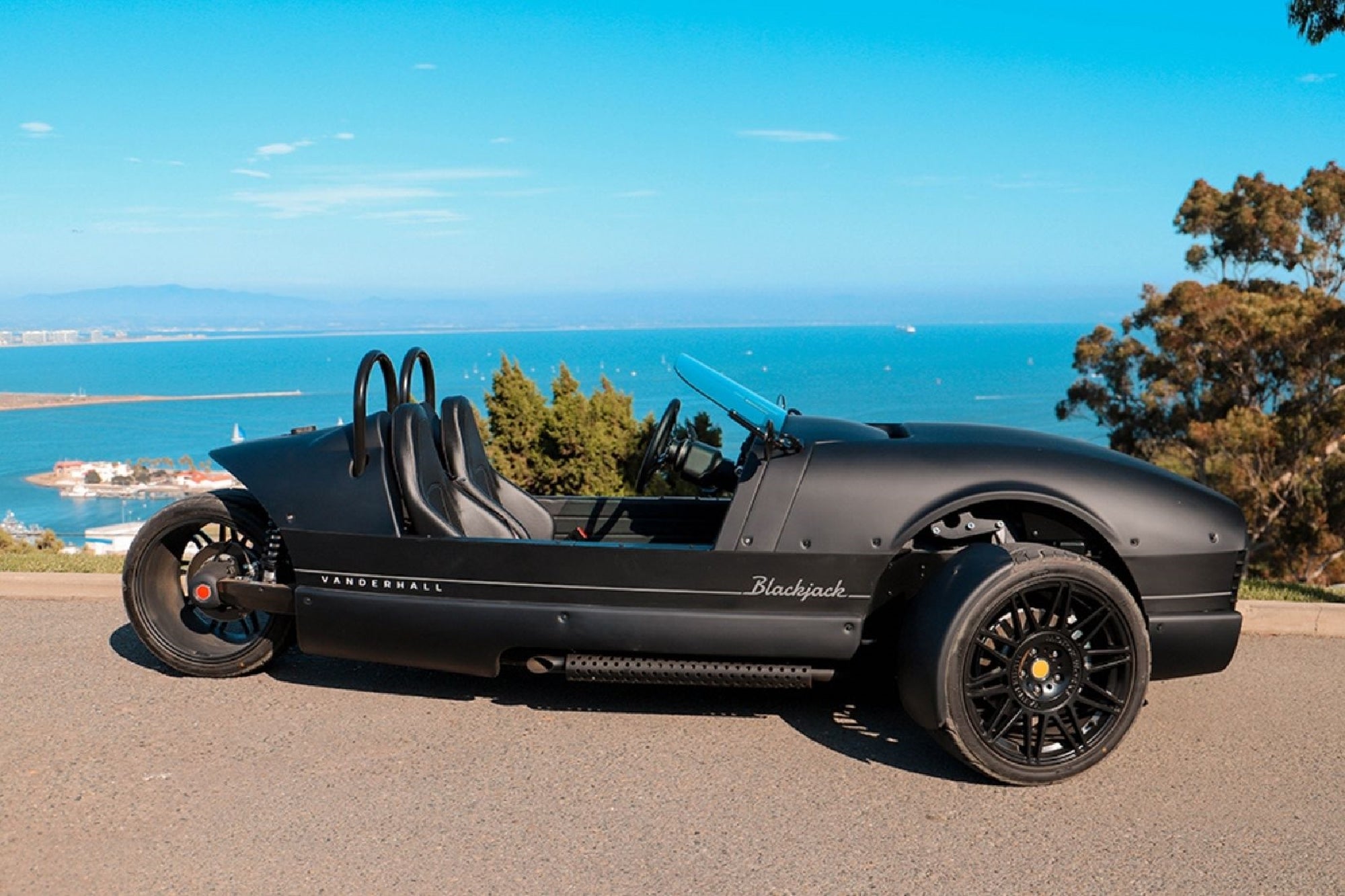A matte black Vanderhall Blackjack three-wheeled roadster is parked on a paved road overlooking the ocean and coastline on a clear, sunny day.
