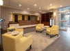 A hotel lobby with yellow armchairs, round tables, a rug, and a front desk in the background. Glass entry doors and shelves are visible to the right.