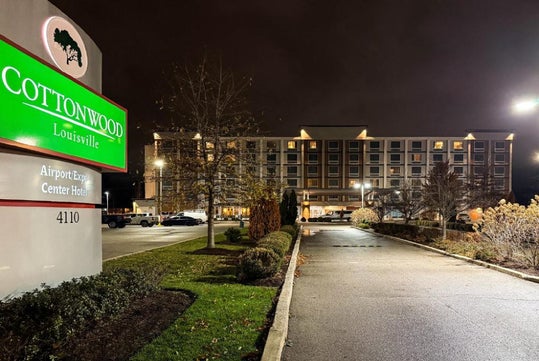 A nighttime view of the Cottonwood Louisville Airport Expo Center Hotel, with a well-lit sign and the hotel building in the background.