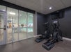 Two elliptical machines are positioned in a small fitness room with a wall-mounted TV and large windows overlooking an indoor swimming pool area.