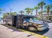 A black tour bus labeled "Count's Kustoms" is parked on a sunny street lined with palm trees, with its door open and cranes visible in the background.