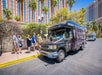 People stand in line to board a black shuttle bus parked on a sunny street, with tall hotels and palm trees in the background.