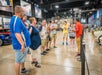 A group of people listens to a guide giving a tour inside a car museum, with classic and modern cars displayed in the background.