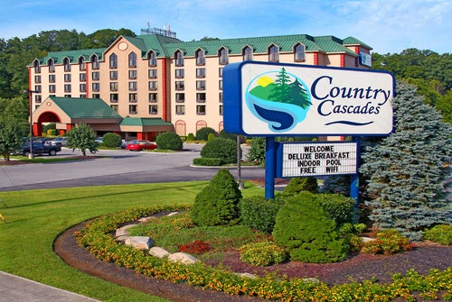 Hotel exterior with a sign reading "Country Cascades." The sign advertises a deluxe breakfast, indoor pool, free WiFi. The building is surrounded by landscaped greenery.