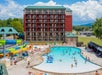 Outdoor pool area with people swimming and lounging, a small waterslide, and a multi-story hotel building in the background on a sunny day.