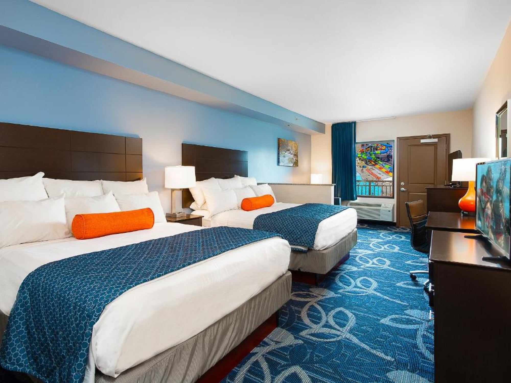 Hotel room with two double beds, each with blue bedding and an orange cushion. A TV, desk, and lamp are visible. Artwork and a window with curtains adorn the space.