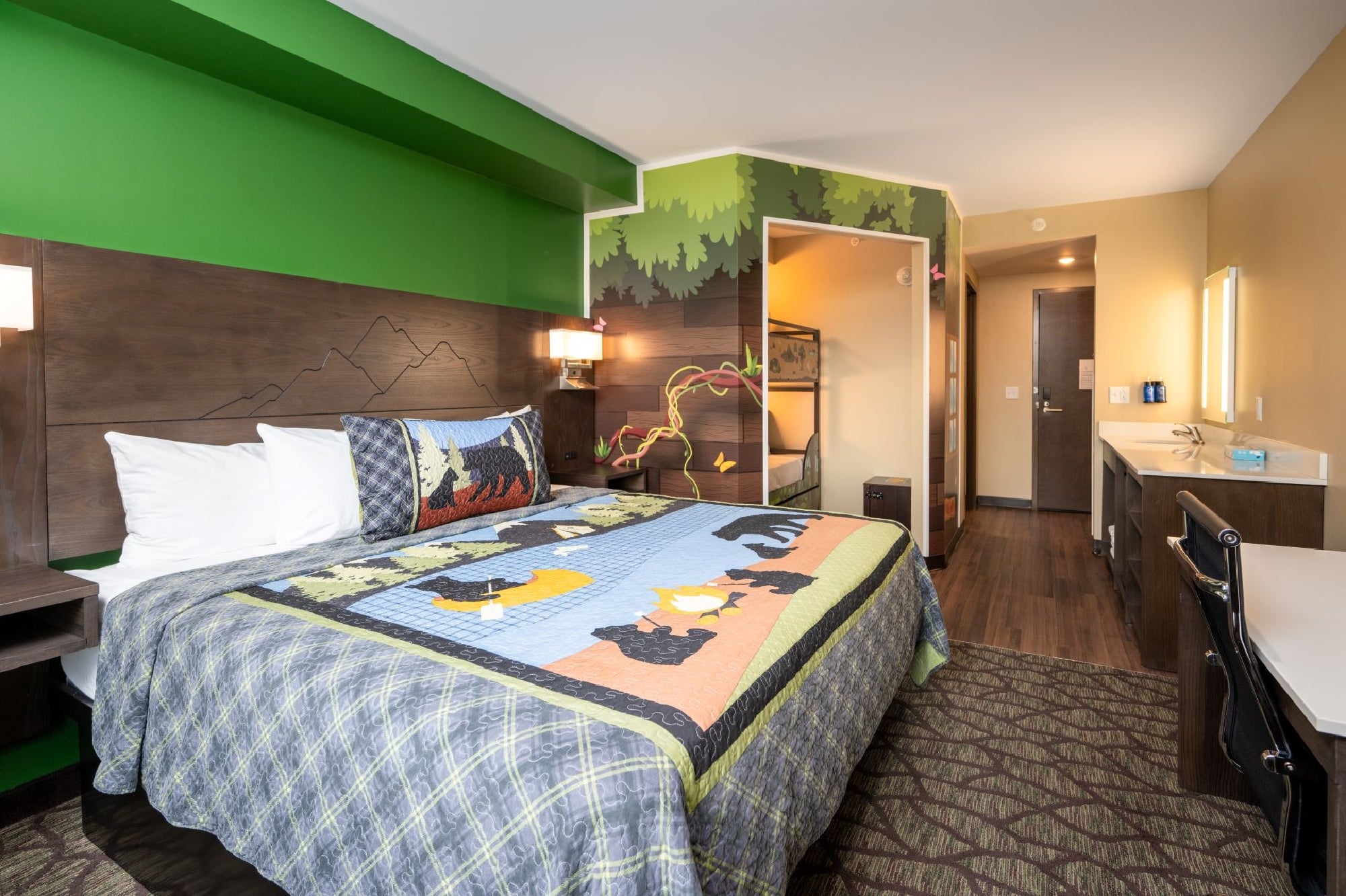 Hotel room with a bed featuring bear-themed bedding, a bunk bed, forest-themed wall art, a desk, a sink area, and a door leading to the hallway.
