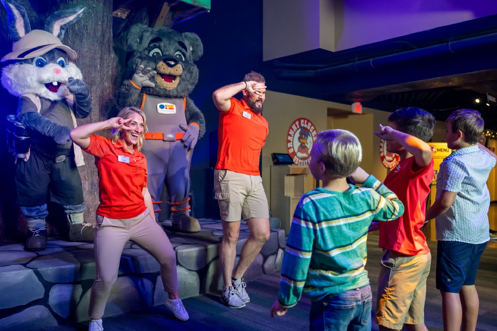 Two adults and several children stand indoors saluting, with two people in animal costumes near them, all participating in an organized group activity.