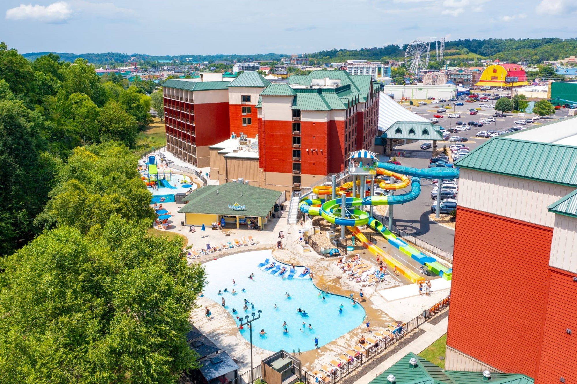 Aerial view of a hotel complex with outdoor swimming pools, water slides, and people swimming; parking lot and commercial buildings are visible in the background.