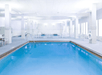 An indoor swimming pool with a smaller pool, hot tub, and white chairs and tables behind it and a wall of windows to the right.