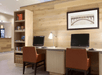 A business center with two computers at a desk with two brown chairs, a shelf full of books to the left and a piece of art on the wall.