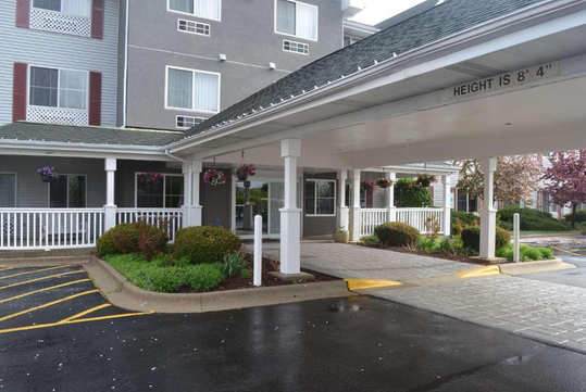 The image shows a hotel entrance with a covered driveway. The sign indicates a height limit of 8 feet 4 inches. Flower pots hang from the ceiling, and there are shrubs and parking lines nearby.