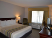Hotel room with a king-size bed, striped bedding, a nightstand with a lamp, an armchair, a window with curtains, an air conditioning unit, and a TV on the dresser.