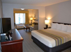 Hotel room with a king-sized bed, sofa, table, and flat-screen TV. Soft lighting and a large window with curtains brighten the space.