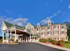 A large country-style hotel with a covered entrance, green roof, and beige siding, surrounded by neatly trimmed shrubbery and a clear blue sky overhead.