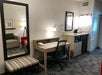 Hotel room with a desk and chair, a large mirror, a striped bench, and a kitchenette area with a sink and mini fridge. A lamp and a phone are on the desk. There's a partial view of a sitting area.