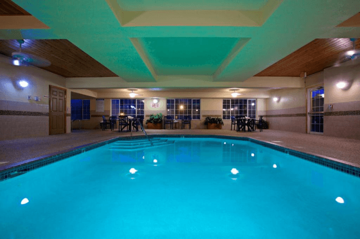 A bright blue large indoor swimming pool with chairs and table on the far end at night.