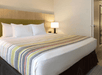 A hotel room with a white king bed with a light wooden headboard and a table and lamp next to it.