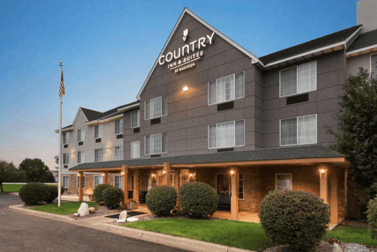 The front exterior of the Country Inn & Suites with several bushes and a flag pole at dusk in Minneapolis, Minnesota.
