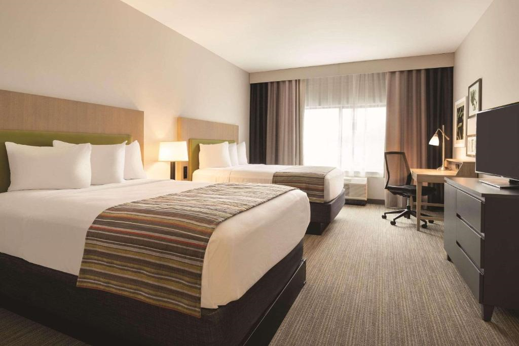 Hotel room with two double beds, a desk with chair, a TV on a dresser, a floor lamp, and large window with curtains. Neutral color palette with light walls and carpet.